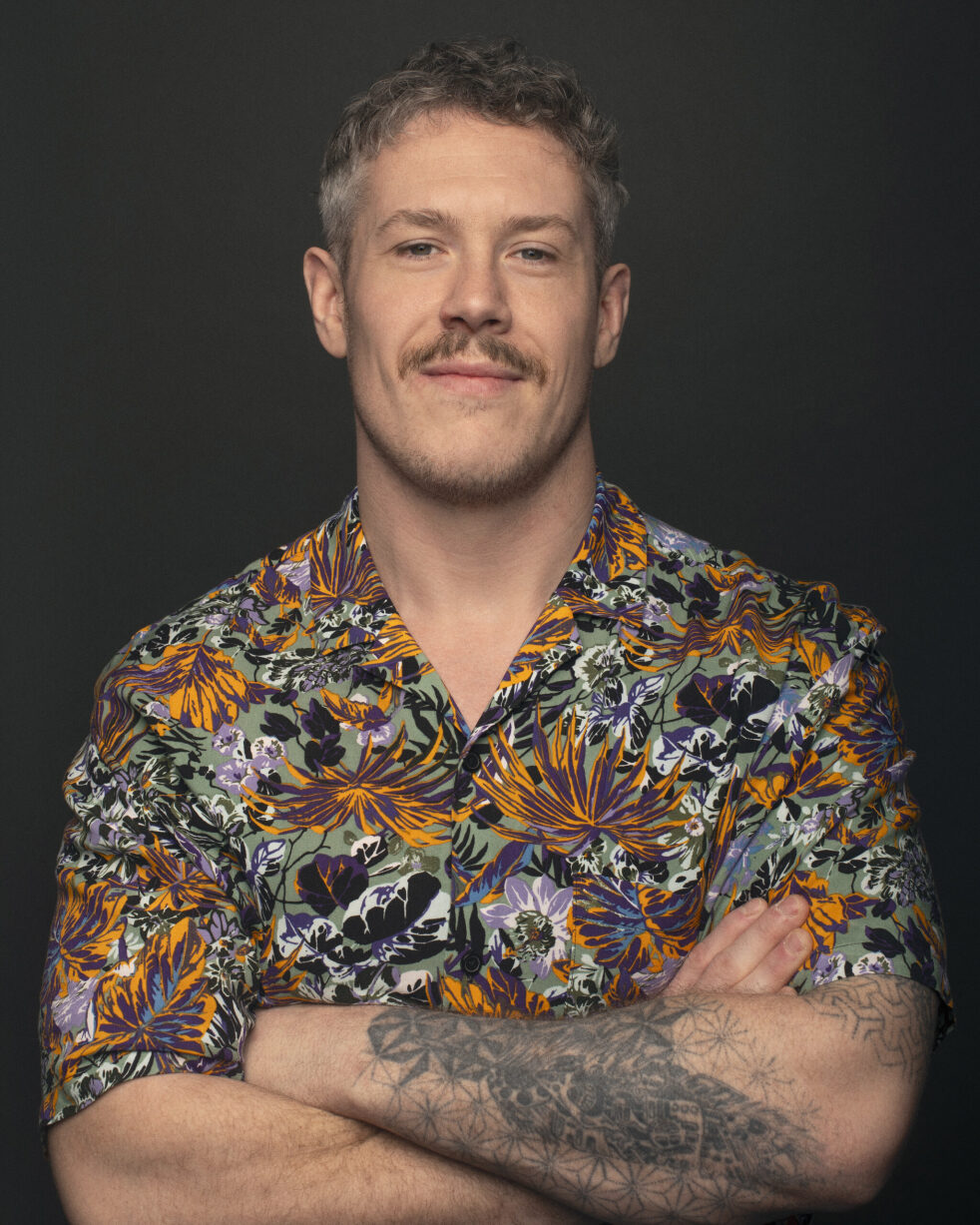 Man with a mustache and a tattooed arm crosses his arms, wearing a vibrant floral shirt. Dark background; confident expression. Alex Beddows headshot.