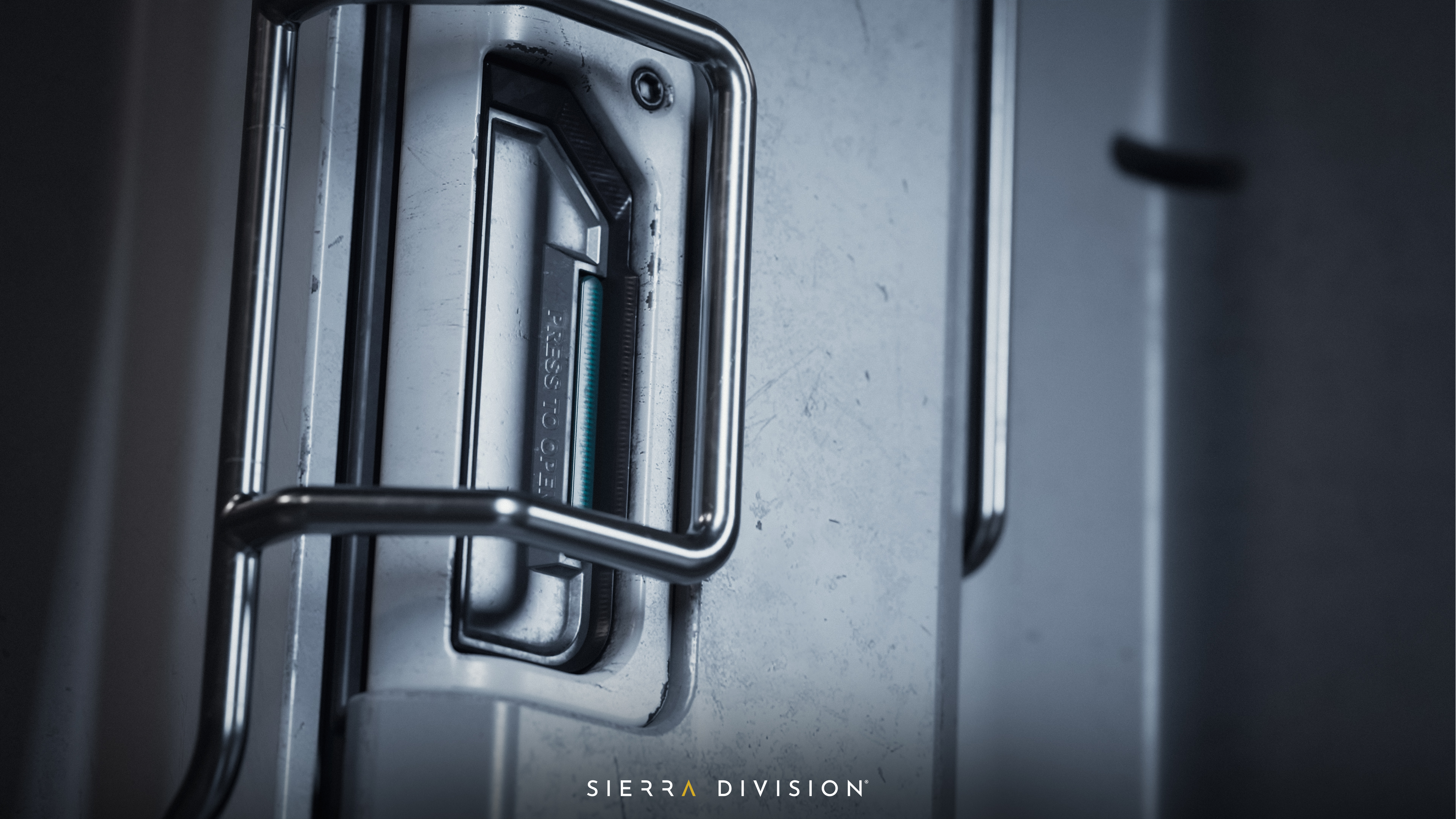 Metal door handle with "PRESS TO OPEN" text, surrounded by a metal frame on a gray surface. Logo "SIERRA DIVISION" below. Industrial feel.