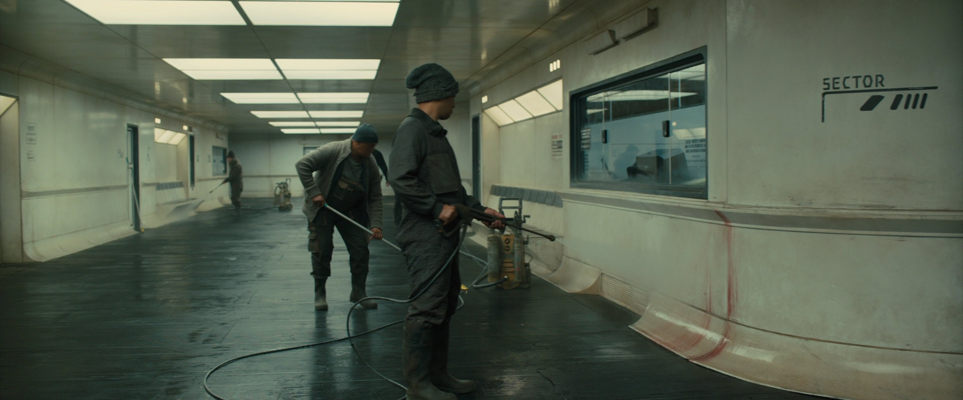 Workers in a dimly lit corridor clean with hoses. Walls show "SECTOR" sign. Floor is wet, and ambiance is industrial. From Blade Runner 2049
