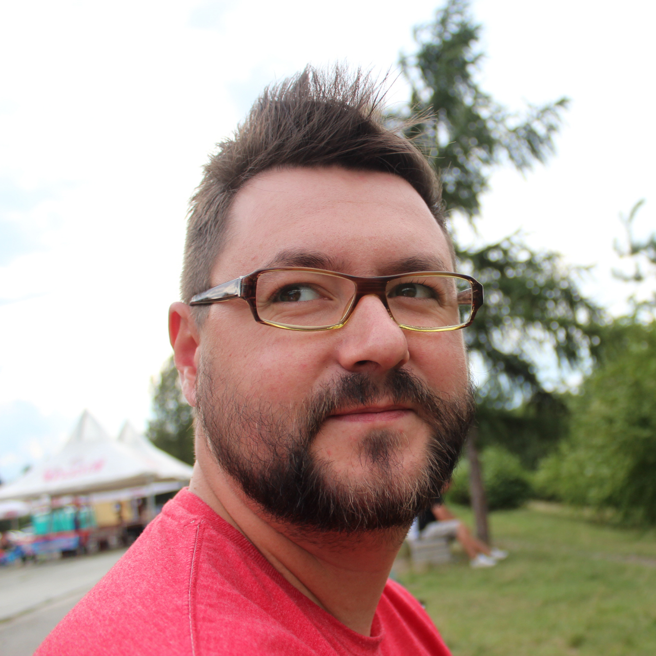 Man with glasses in a red shirt smiles outdoors, surrounded by trees and tents. The setting feels casual and relaxed.