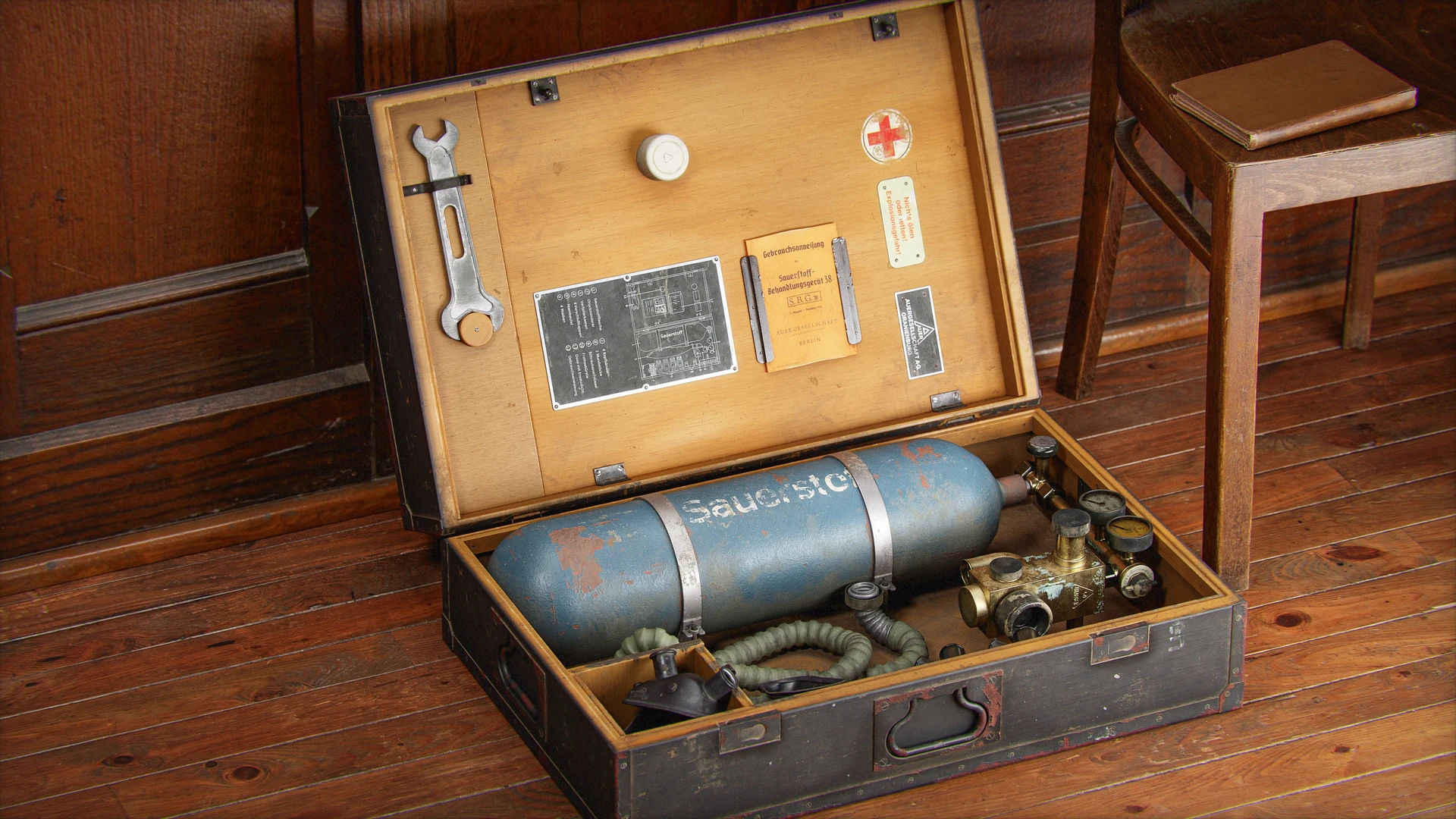 Open wooden boxes on a wooden floor reveal coiled hoses, a blue cylinder with "Sau" text, gauges, and tools. A canvas cover and shoes visible.