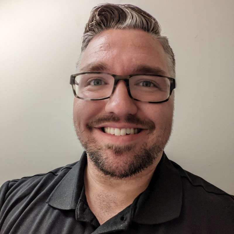 Headshot of Gregory Robb. Smiling person with glasses and a beard, wearing a dark shirt against a plain, light background. Friendly and relaxed mood.