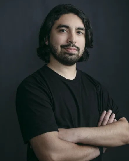 Headshot of Jon Arellano. Long hair and beard in a black shirt stands confidently with crossed arms against a dark background, calm expression.