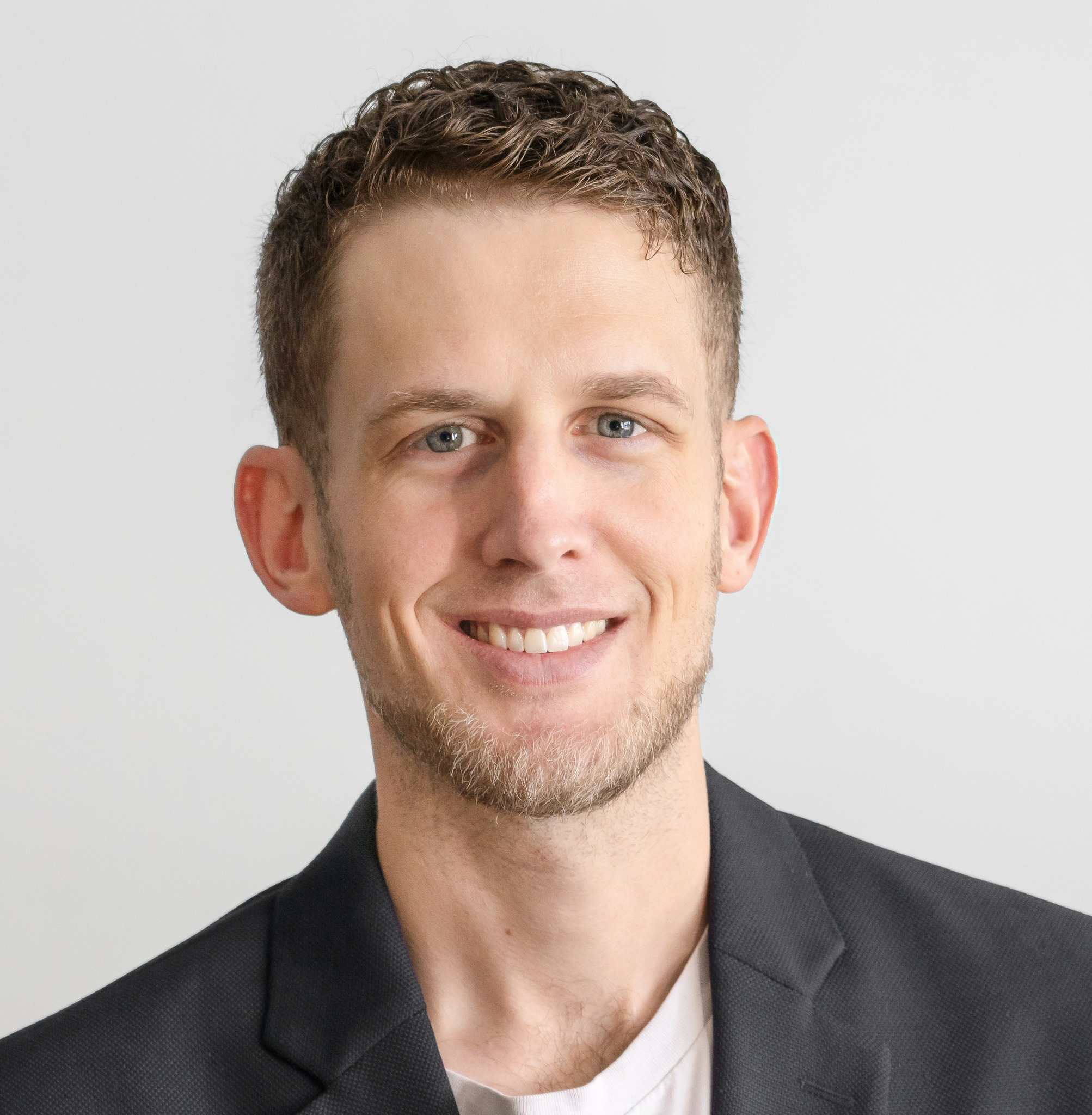 Headshot picture of Jacob Norris. Smiling man with short wavu hair in a black blazer and white shirt. Neutral gray background, conveying a professional, friendly mood.